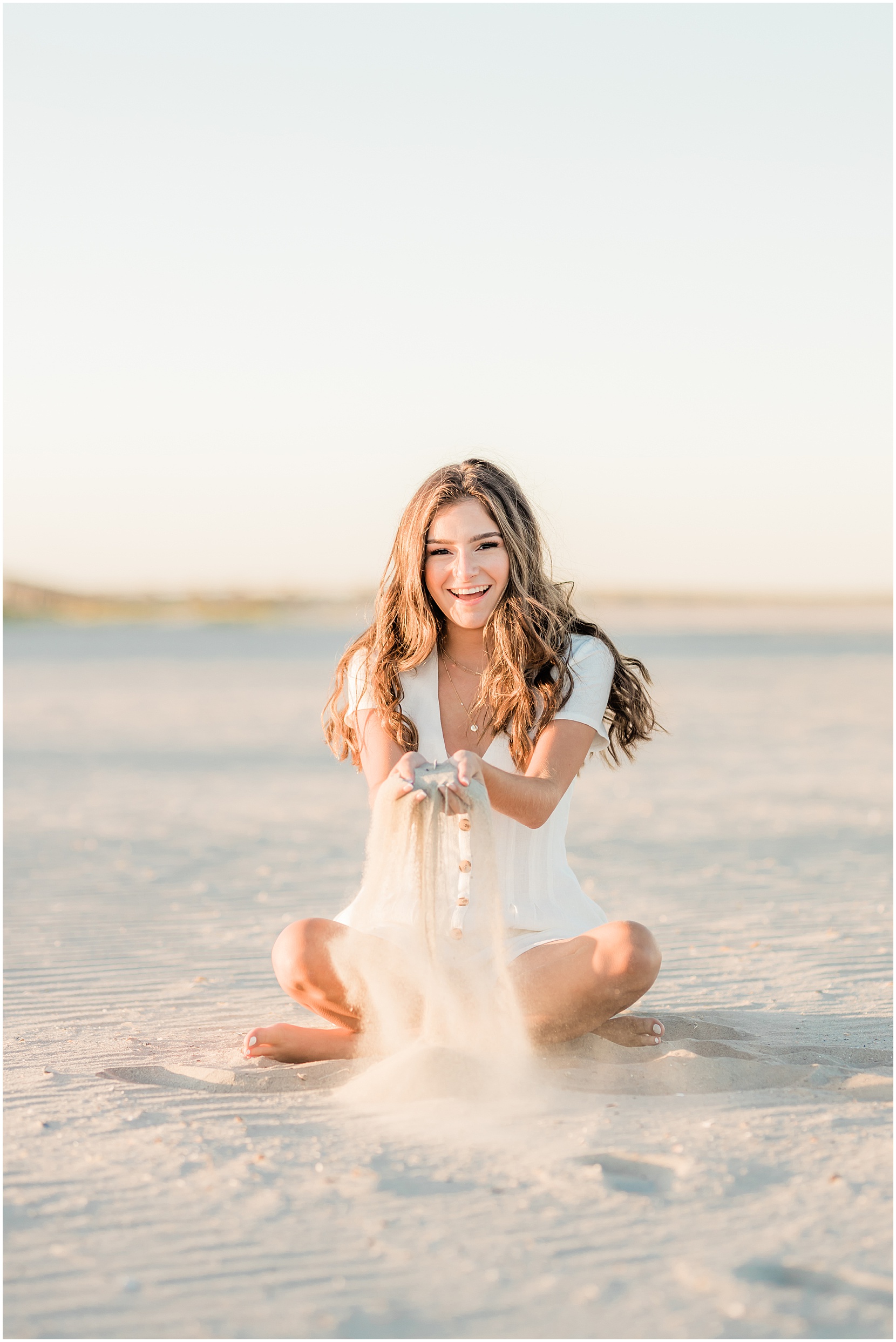 High school senior girl posing for senior pictures on the beach in ocean city new jersey. she is wearing a white romper and she is playing in the sand. the sun is golden!
