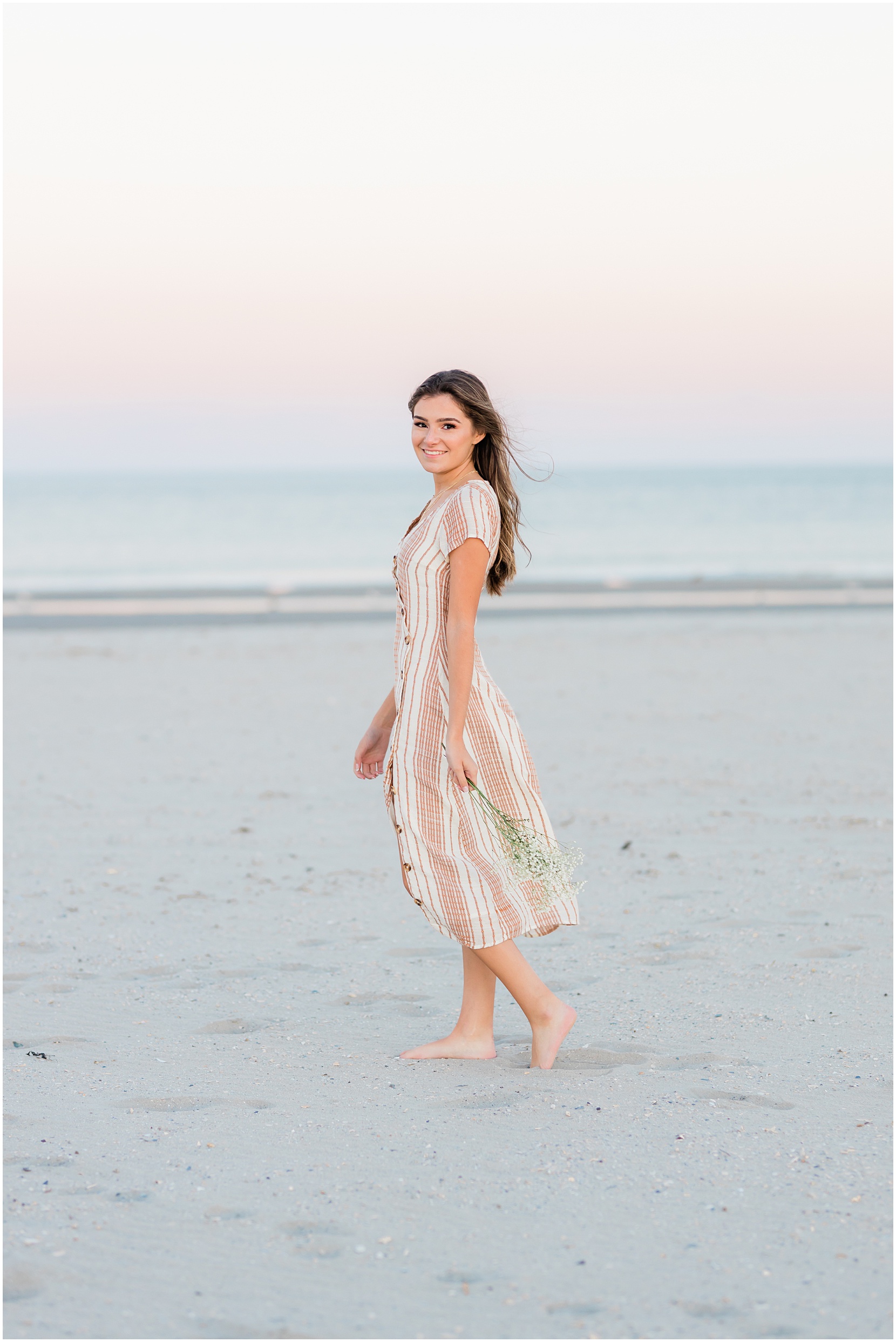 Teen girl posing for her senior pictures with Nicole Marie Photography. She is standing in the under the pink sunset sky in Ocean City New Jersey.