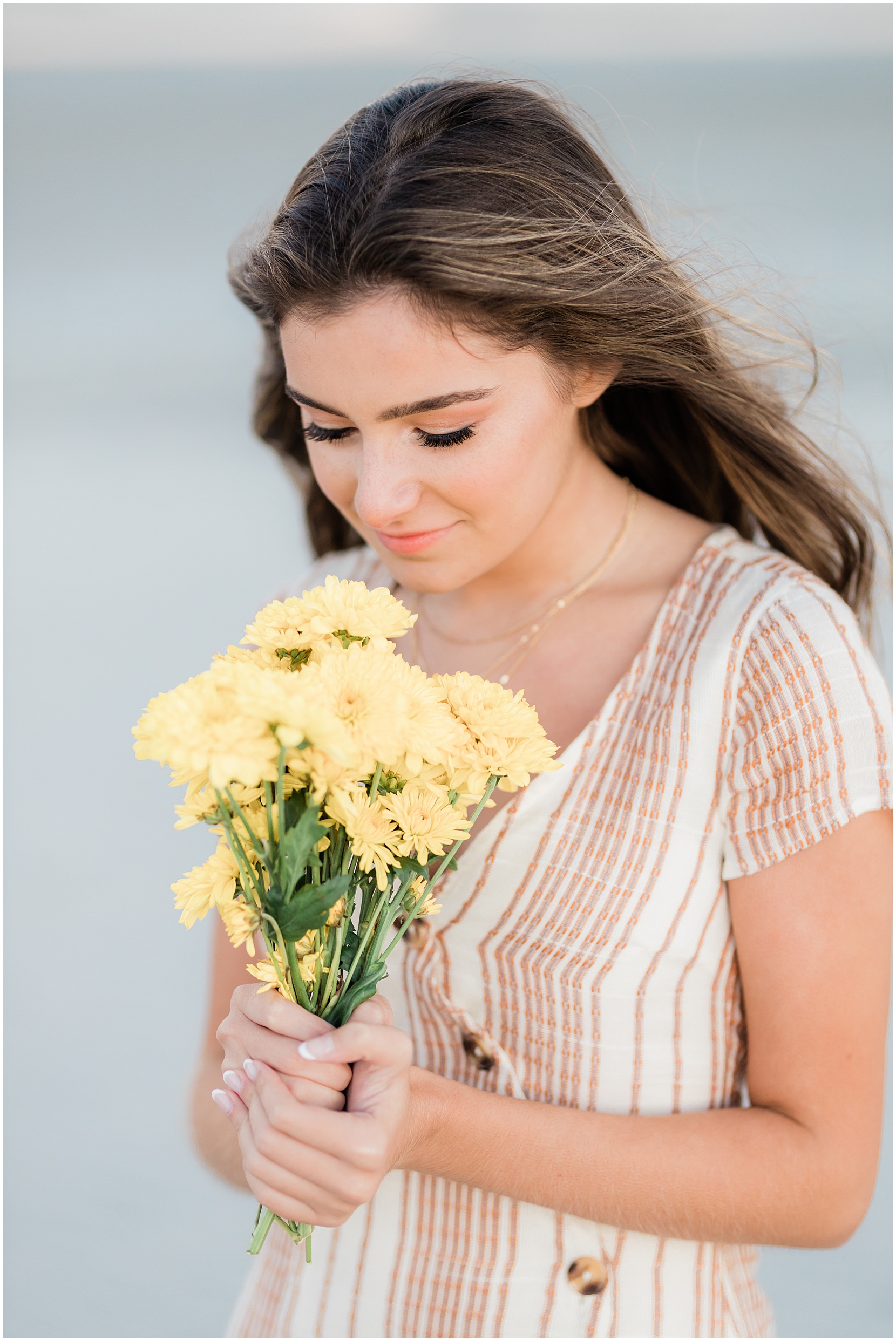 Teen girl posing for her senior pictures with Nicole Marie Photography. She is standing in the under the pink sunset sky in Ocean City New Jersey. She is holding yellow daisies.