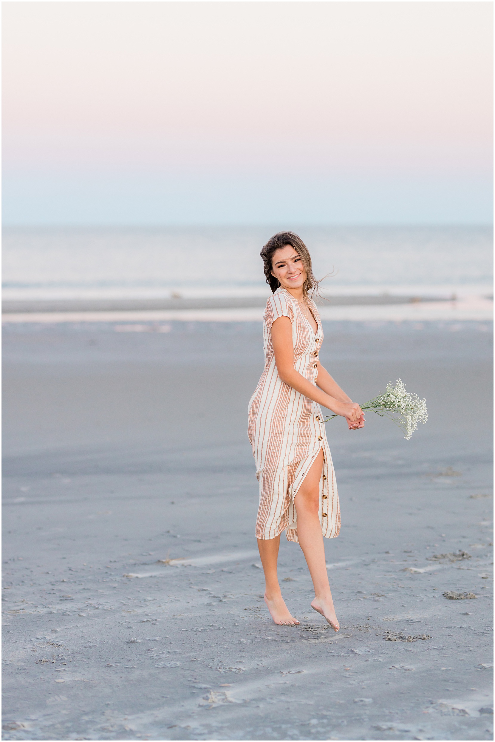 Teen girl posing for her senior pictures with Nicole Marie Photography. She is standing in the under the pink sunset sky in Ocean City New Jersey. She is dancing holding white flowers.