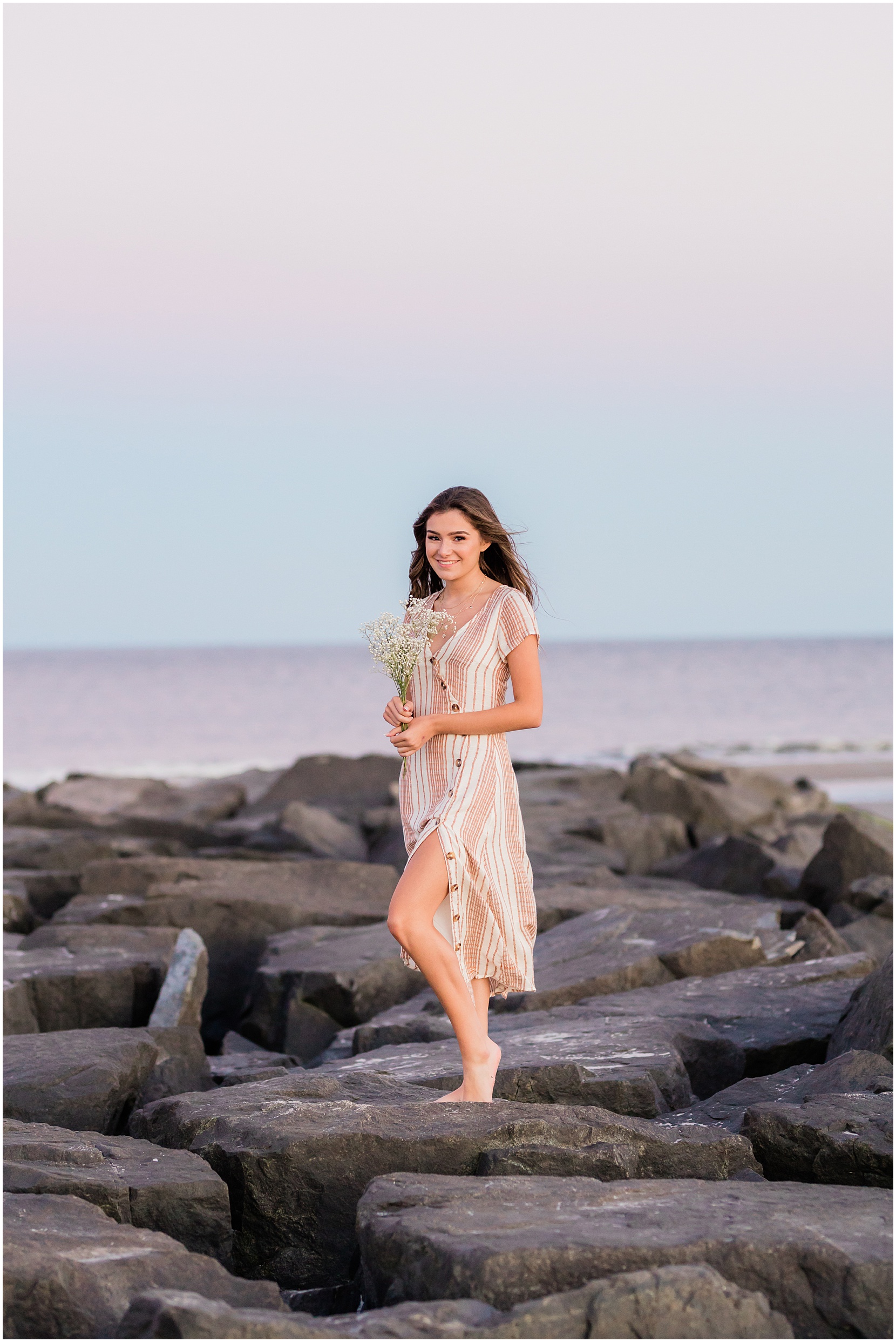 Teen girl posing for her senior pictures with Nicole Marie Photography. She is standing in the under the pink sunset sky in Ocean City New Jersey. She is on the jetty.