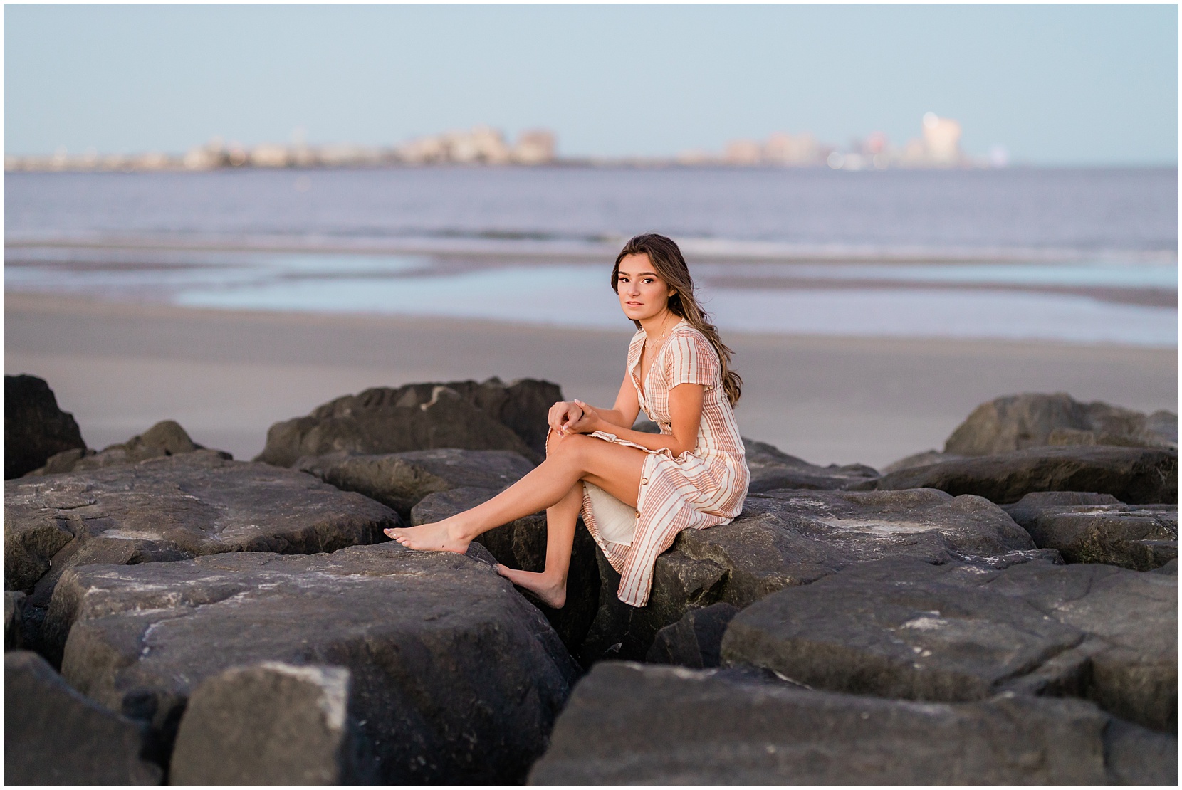 Teen girl posing for her senior pictures with Nicole Marie Photography. She is standing in the under the pink sunset sky in Ocean City New Jersey. She is sitting on the jetty in a tan dress.