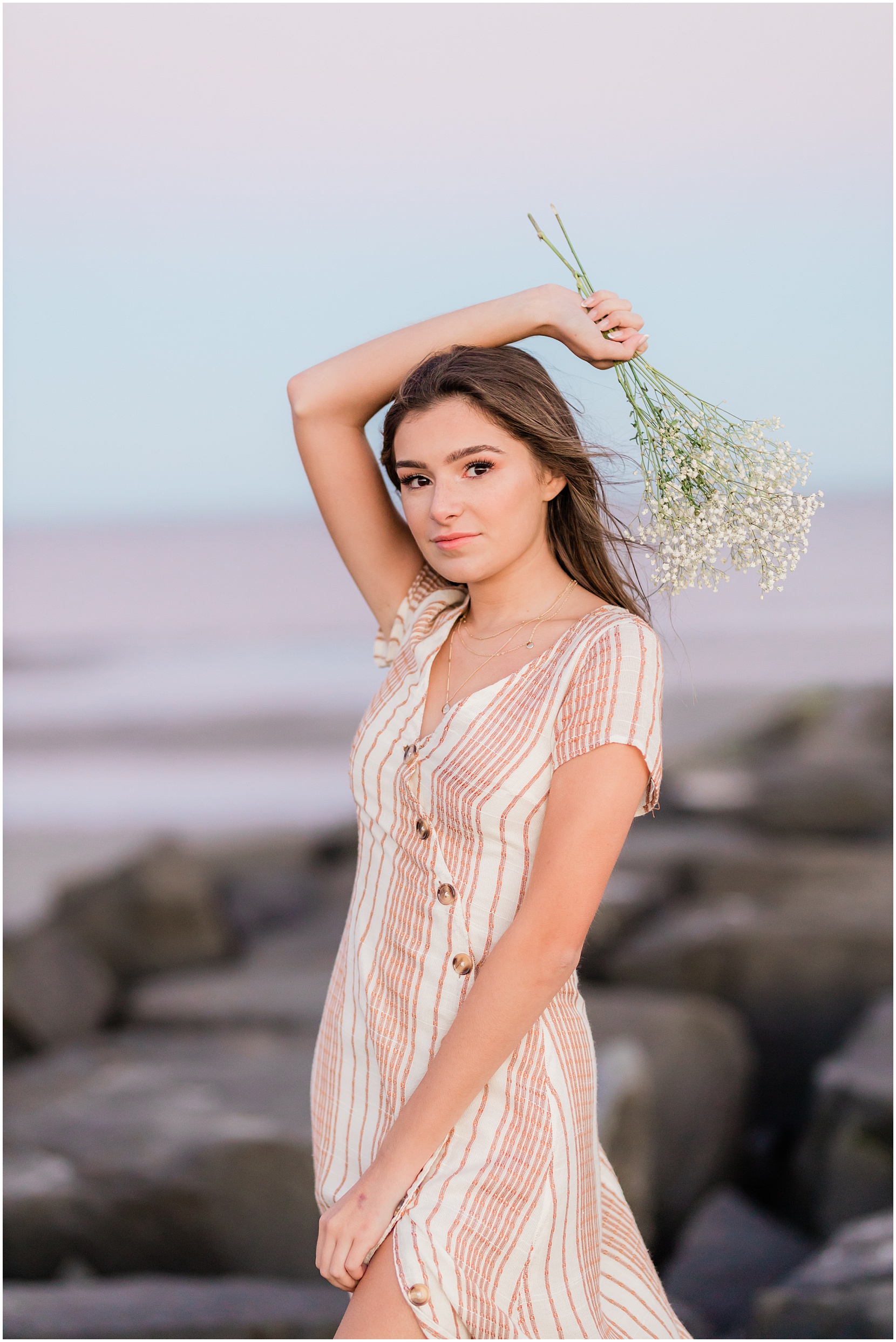Teen girl posing for her senior pictures with Nicole Marie Photography. She is standing in the under the pink sunset sky in Ocean City New Jersey. She is holding white flowers.